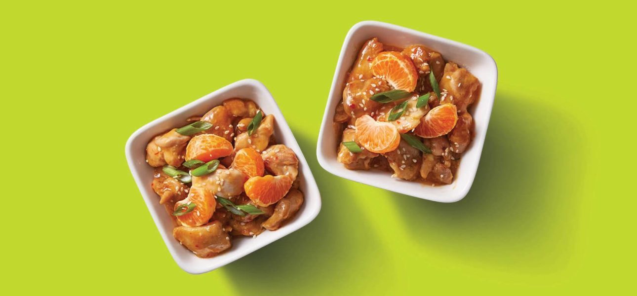 Two white bowls containining mandarin orange chicken, served with orange sauce, orange slices, spring onion, and sesame seeds.